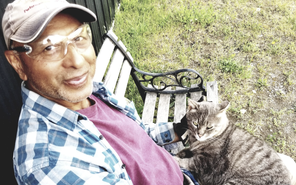 Caregiver Mo sits on a bench with his ear-tipped grey barn cat in his lap. He's smiling at the camera while petting Catsby's cheek.